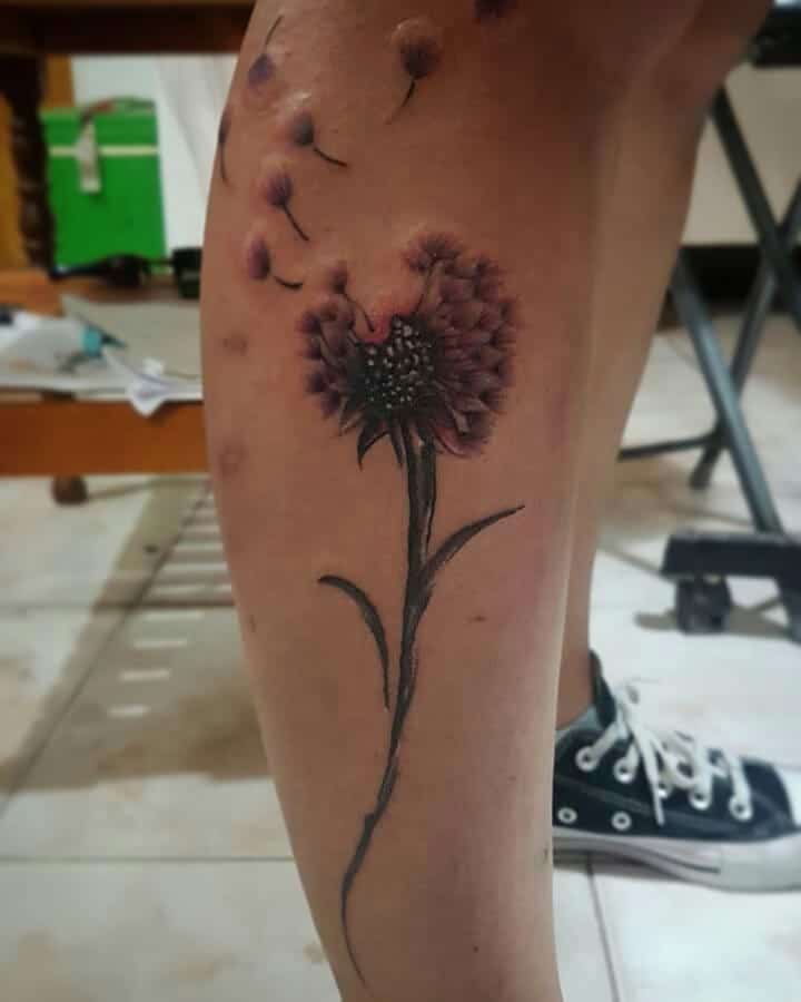 A tattoo of a purple dandelion flower with seeds floating away, placed on the lower leg, with detailed shading and realistic style.