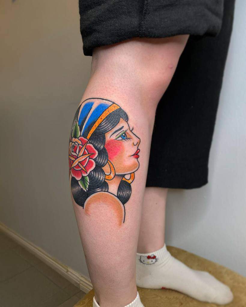 A colorful tattoo on a leg showcases a gypsy woman's profile, adorned with a rose and a striped headscarf