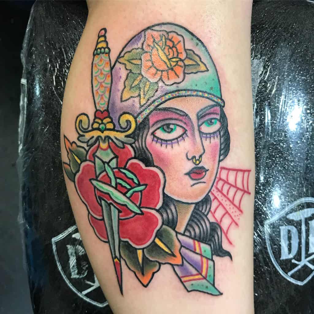 Tattoo of a gypsy woman's face adorned with a helmet, accompanied by a rose, dagger, and web on the arm