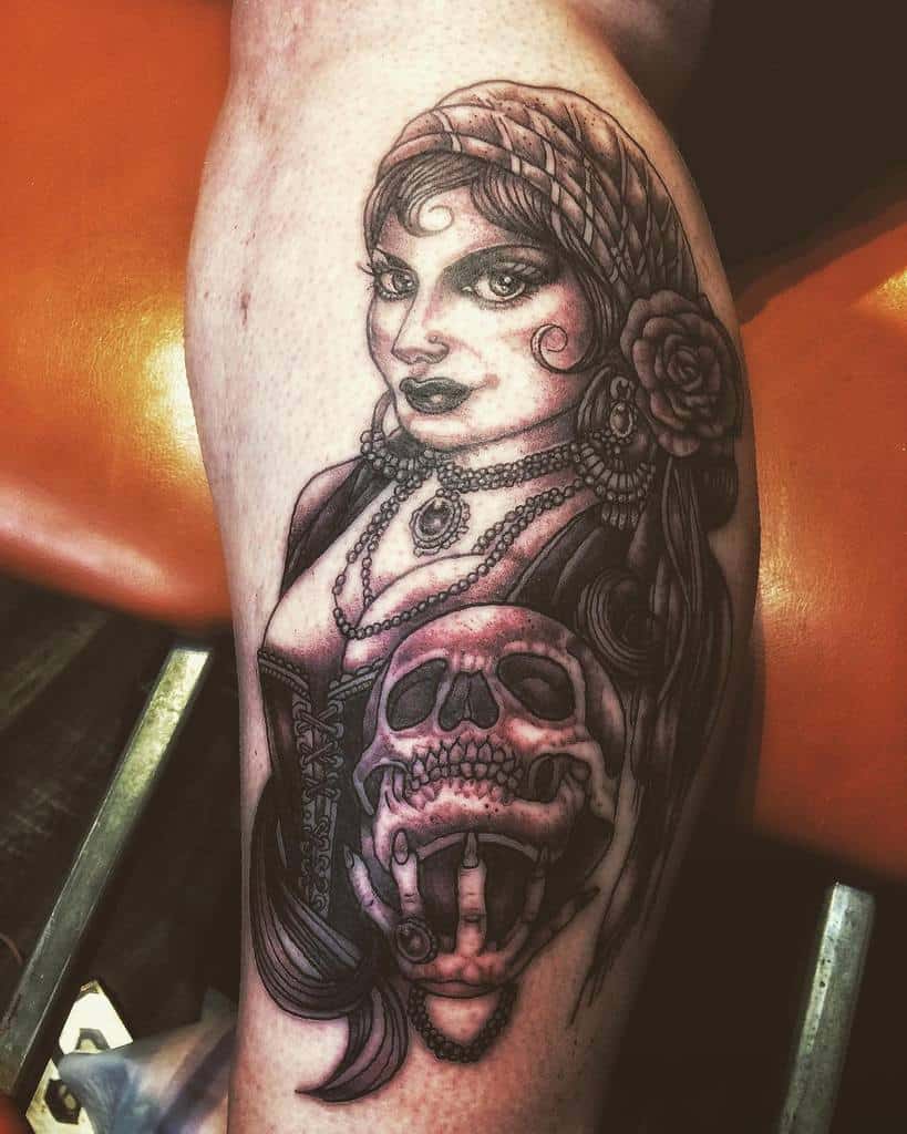 A black and gray tattoo of a gypsy woman with a headscarf and rose, holding a skull with a hand emerging from it, on the leg