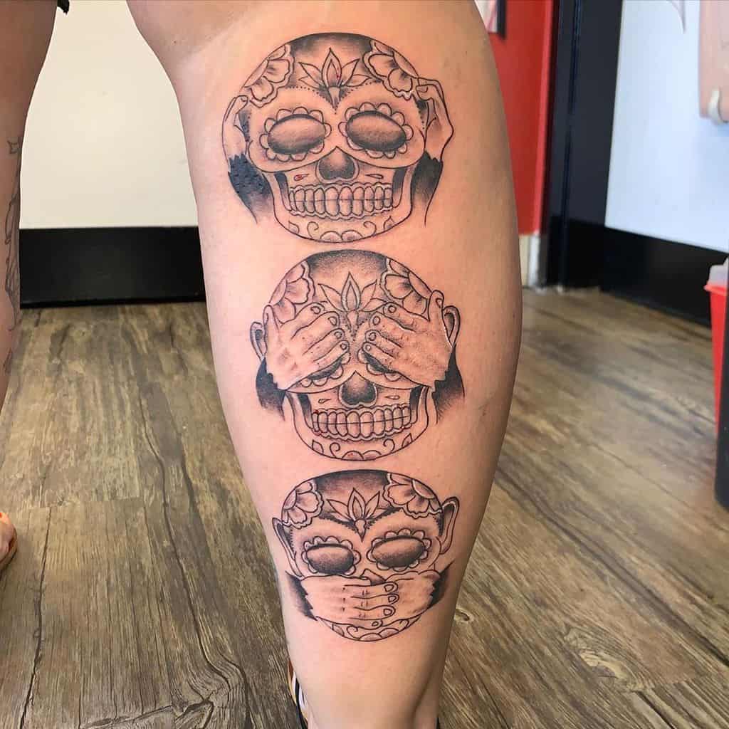 Leg tattoo of three stylized skulls with hands covering eyes, ears, and mouth, depicting "see no evil, hear no evil, speak no evil"