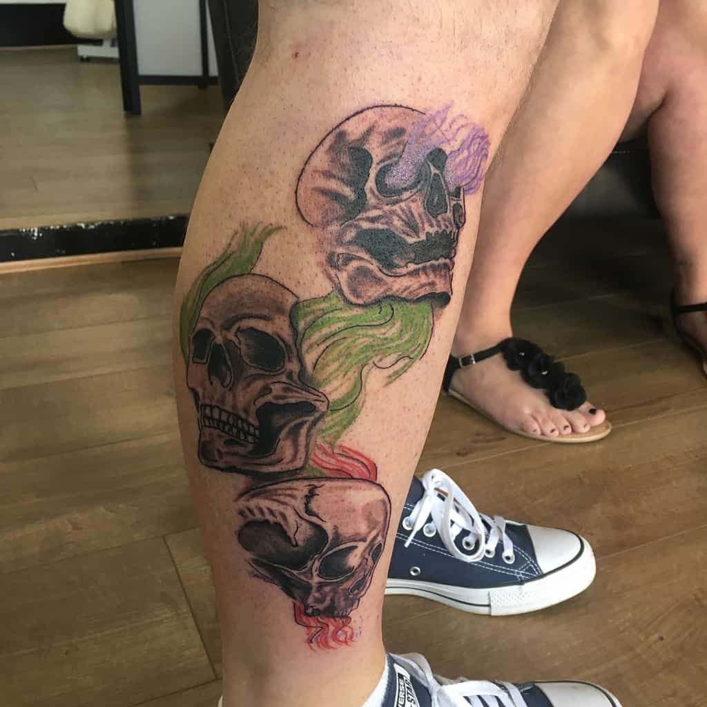 Colorful leg tattoo of three skulls aligned vertically, each representing 'Hear No Evil, See No Evil, Speak No Evil,' with flowing vibrant accents in green, purple, and red