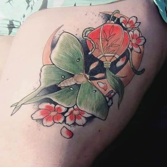 A vibrant tattoo of a Luna Moth surrounded by pink flowers and an orange lantern with a crescent moon behind it