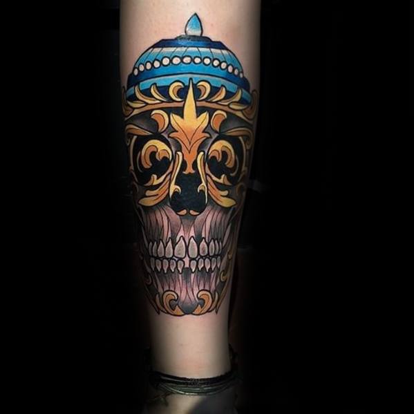 Colorful Tibetan skull tattoo on the calf with blue crown, golden ornamental accents, and detailed teeth on a black background.