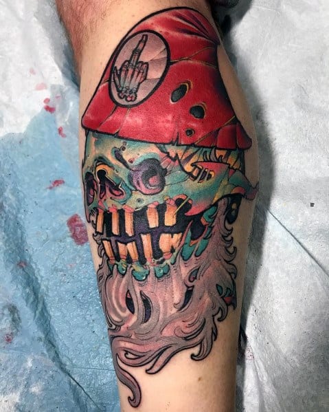 Tattoo of a sinister gnome with a decayed skull face, glowing green eyes, a tattered red hat, and a middle finger patch, blending horror and fantasy.