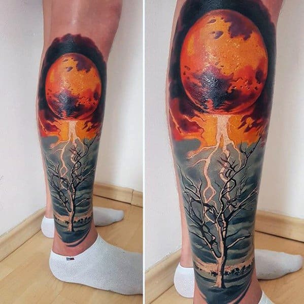 Surreal leg tattoo of a fiery orange moon erupting lightning into a barren tree, set against a dramatic sky and desolate landscape.