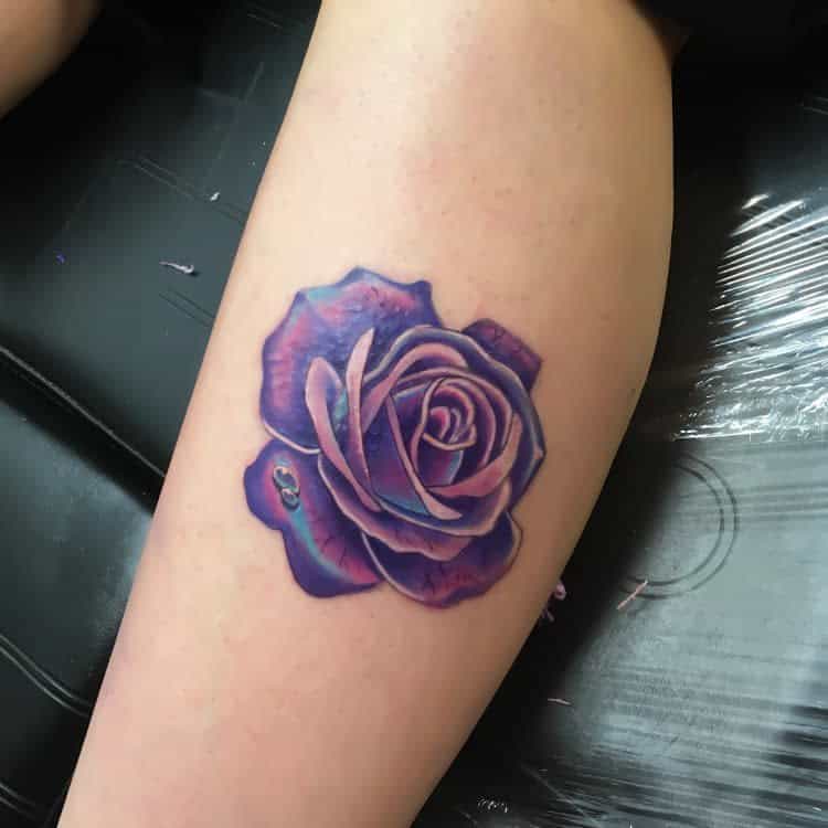 A vibrant tattoo of a purple rose with intricate shading and soft highlights, placed on the lower leg