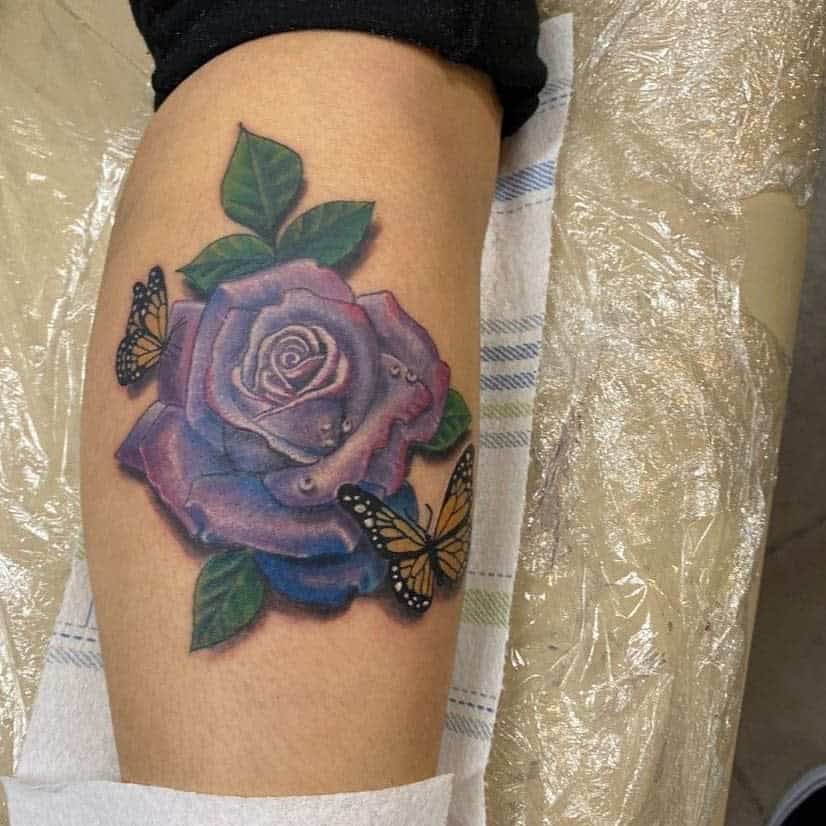 A delicate tattoo of a vibrant purple rose beautifully entwines with fluttering butterflies on an arm