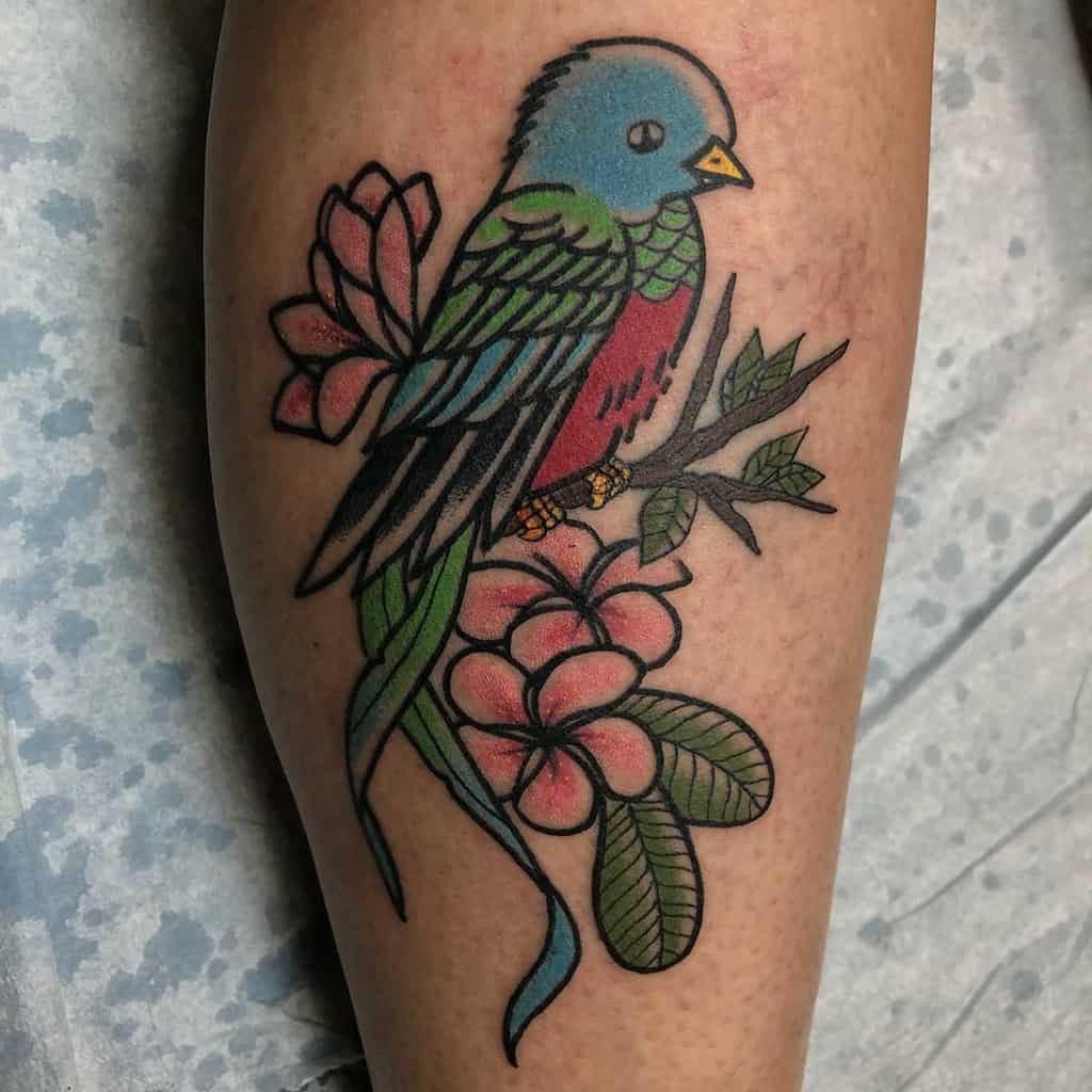 Traditional-style quetzal tattoo on the leg, featuring a colorful blue and red bird perched on a branch with pink flowers and green leaves
