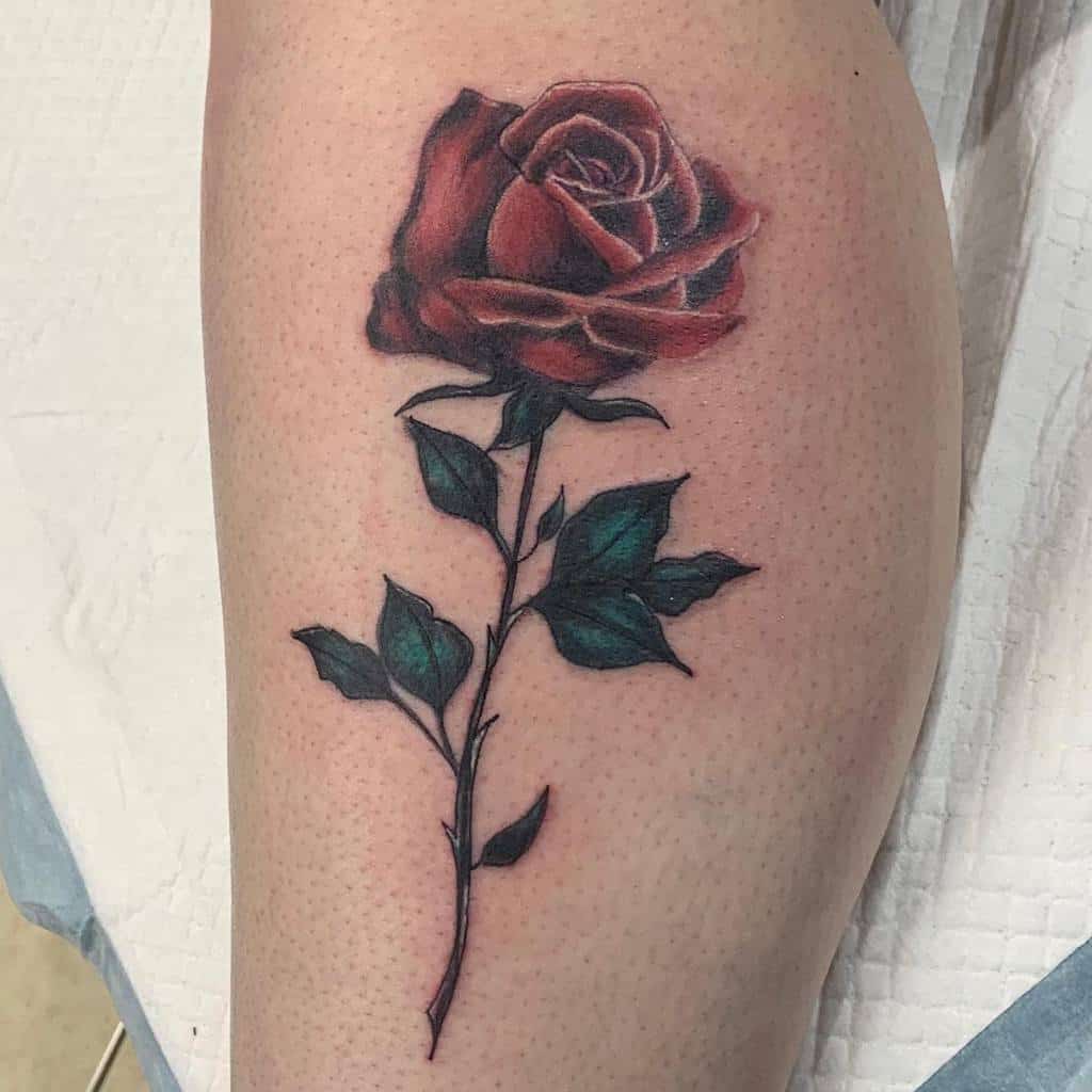 A realistic red rose tattoo on the leg, featuring detailed shading and vibrant colors, with bold outlines that emphasize the delicate petals and leaves