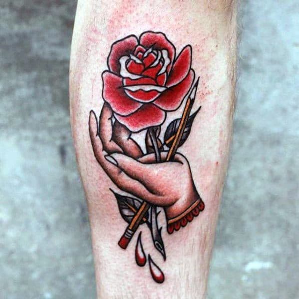 Tattoo of a hand holding a red rose with thorns and a paintbrush, drops of blood below, intertwining delicately with a pencil