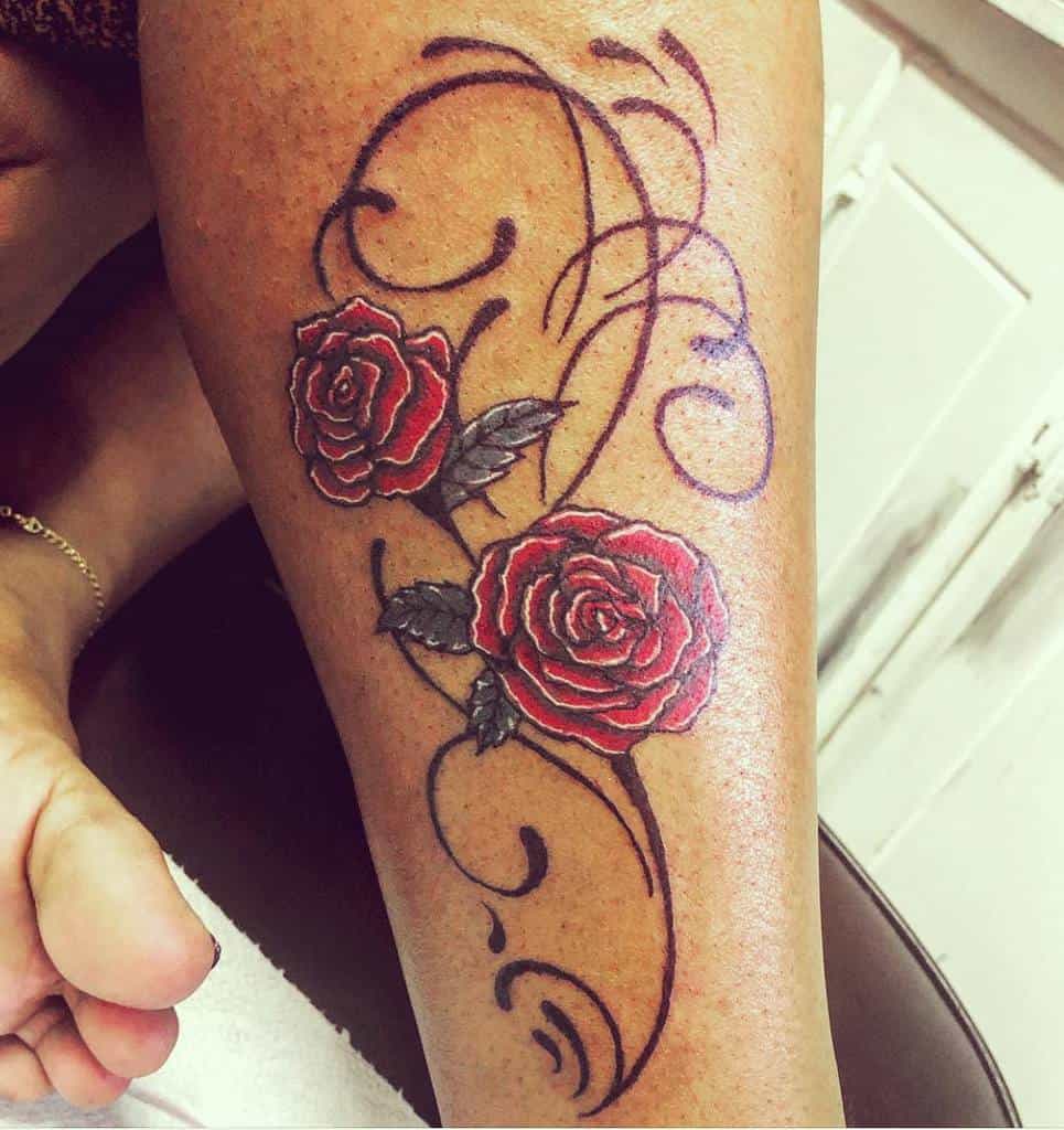 Leg tattoo featuring two bold red roses accented by swirling black vines and green leaves, creating a striking floral design