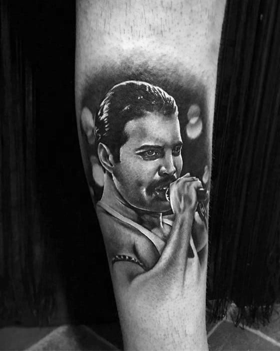 Realistic black and grey tattoo of Freddie Mercury holding a microphone, with soft background lighting and detailed facial features.