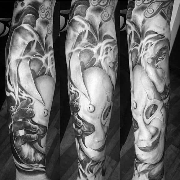 A black and gray tattoo of a jester's face intertwined with abstract swirling smoke and hands, creating a dynamic, eerie design on the forearm.