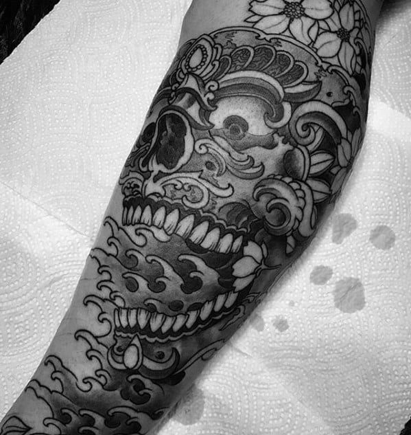 Black and grey Tibetan skull tattoo on leg with wave patterns, floral accents, and intricate ornamental crown details.