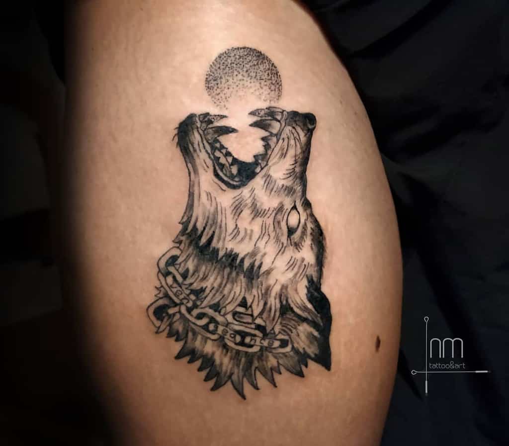 This thigh tattoo features a Norse-inspired wolf with a chain detail, howling at a textured, circular moon—a representation of strength and mysticism