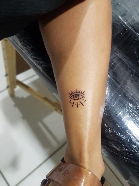 Small minimalist tattoo of an all-seeing eye with radiating lines, inked in black on the lower leg.