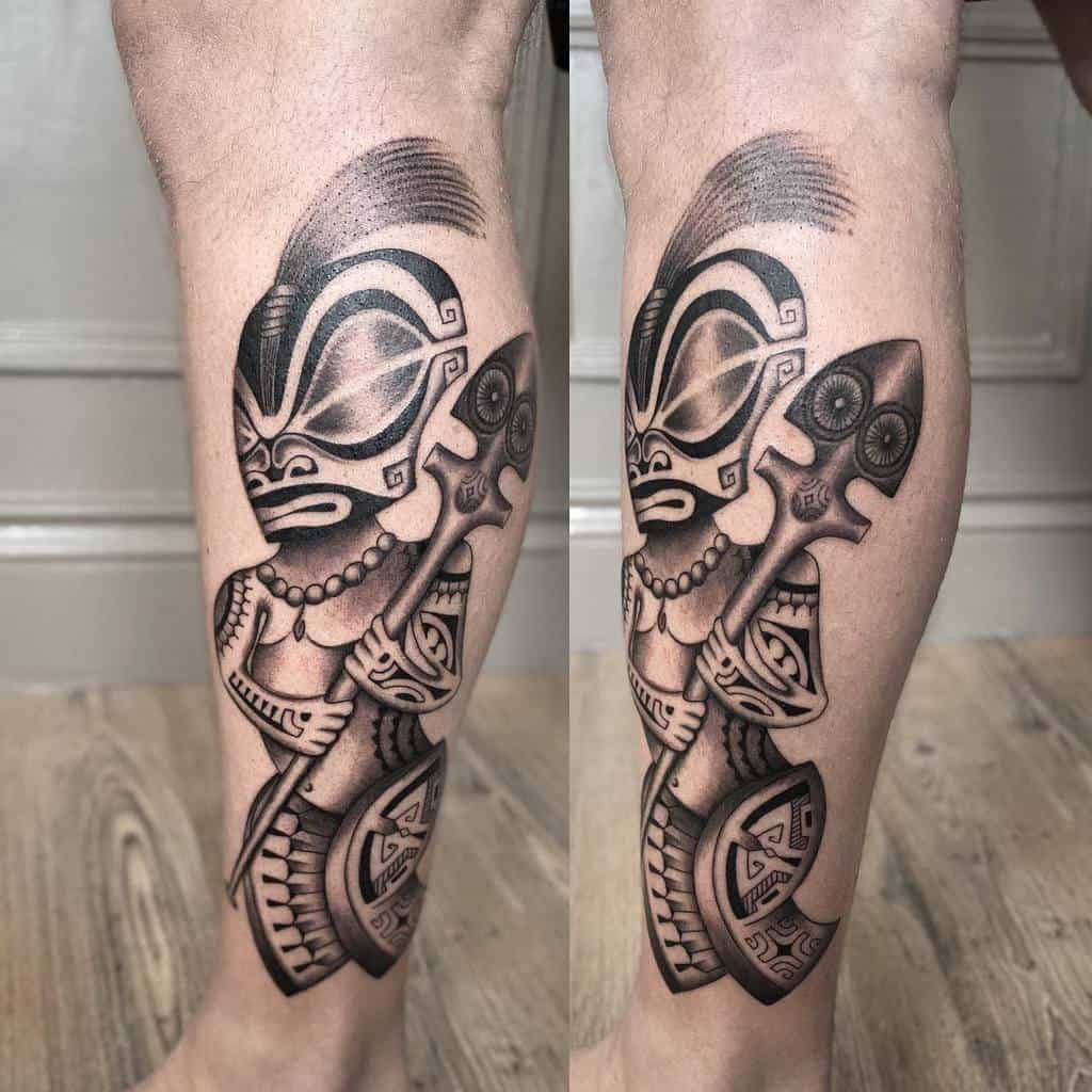 A black-and-gray tattoo of a Tiki mask holding a spear with pearl necklace and adorned with tribal patterns