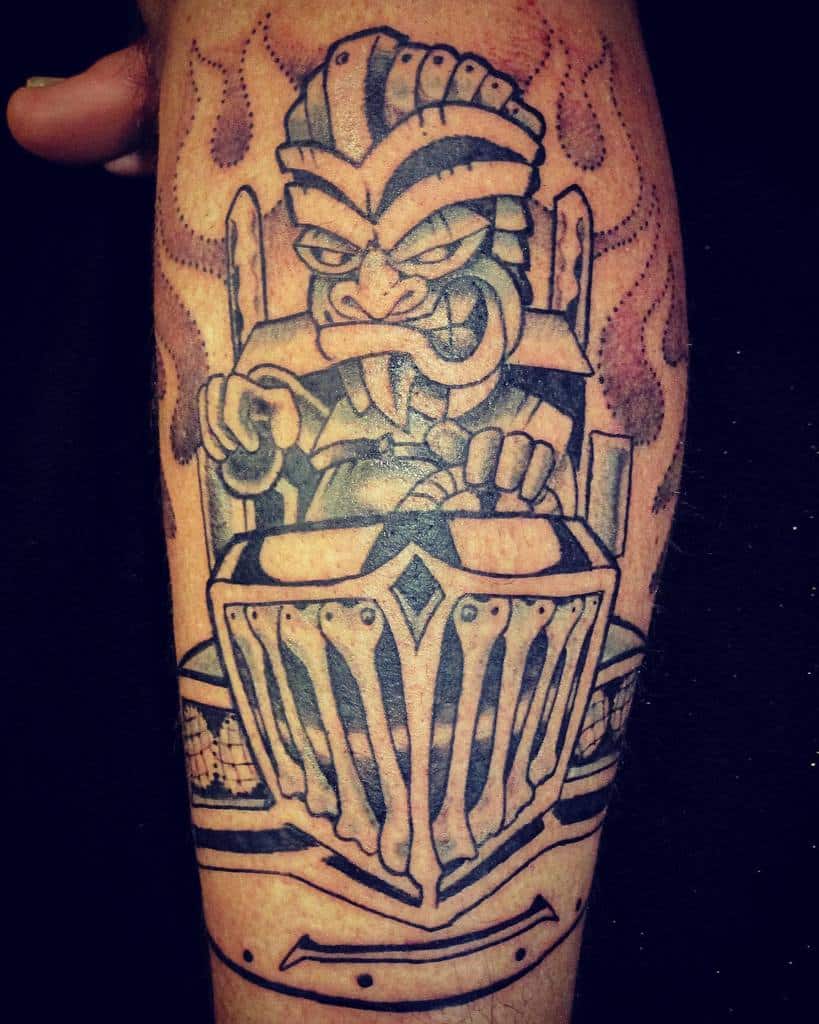 A bold black-and-white tattoo of a Tiki mask with a fierce expression, sitting in a chariot-like vehicle with flames in the background