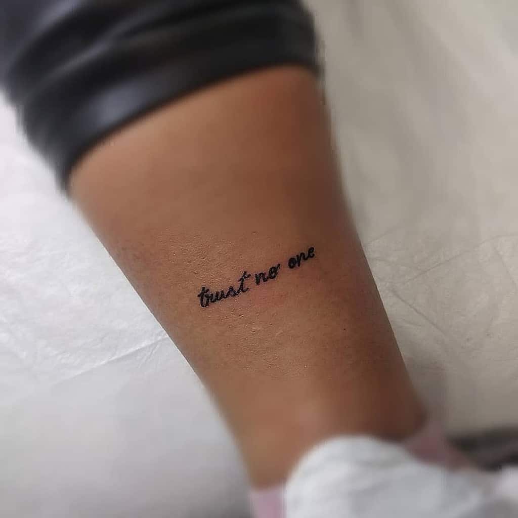 Tattoo on a lower leg reading "trust no one" in cursive writing
