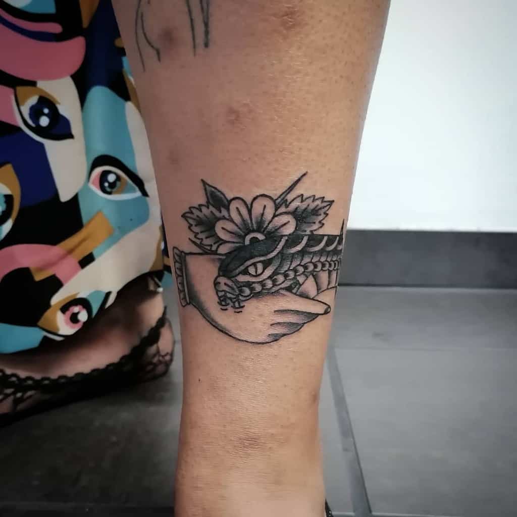 Black ink tattoo of a hand shaking with a snake, adorned with a flower and leaves, placed on the lower leg