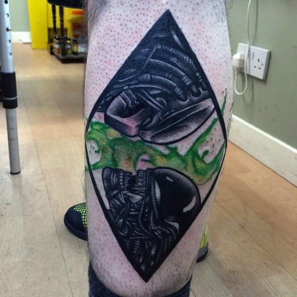 Geometric Xenomorph tattoo in a diamond frame with two alien heads and green smoke-like accents on the calf.