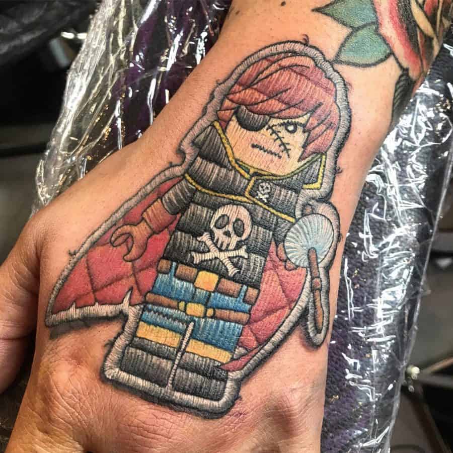 Embroidery-style tattoo of a pirate character on the hand, featuring stitched textures, an eyepatch, a skull emblem, and a cape.