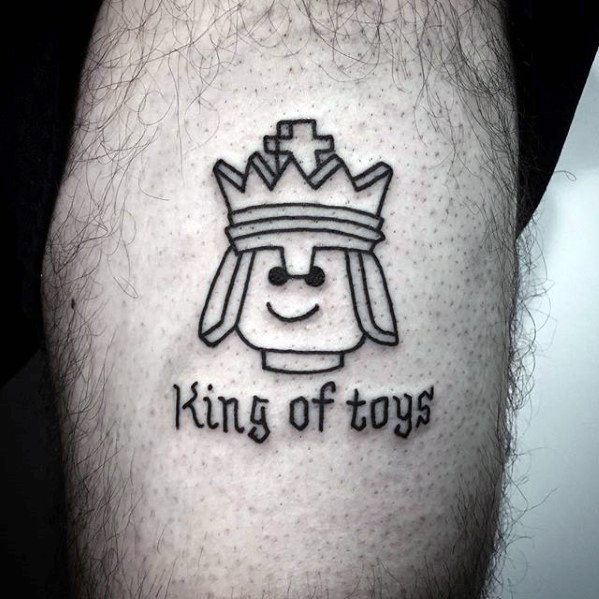 Simple black line tattoo of a Lego king head with a crown, smiling face, and the text "King of toys" in medieval-style font.