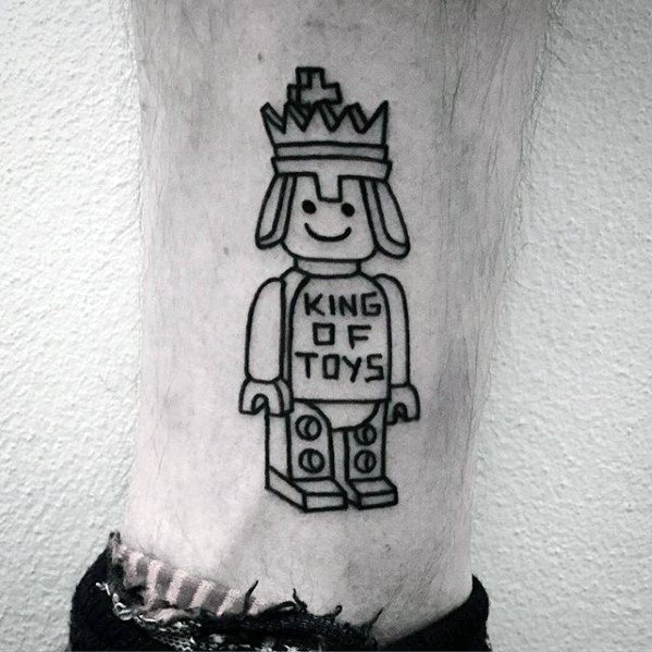 Simple black line tattoo of a full Lego king figure wearing a crown and a shirt that reads "KING OF TOYS," placed on the lower leg.