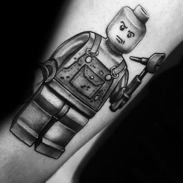 Black and grey tattoo of a Lego mechanic minifigure with a serious expression, wearing overalls and holding a wrench and power drill.