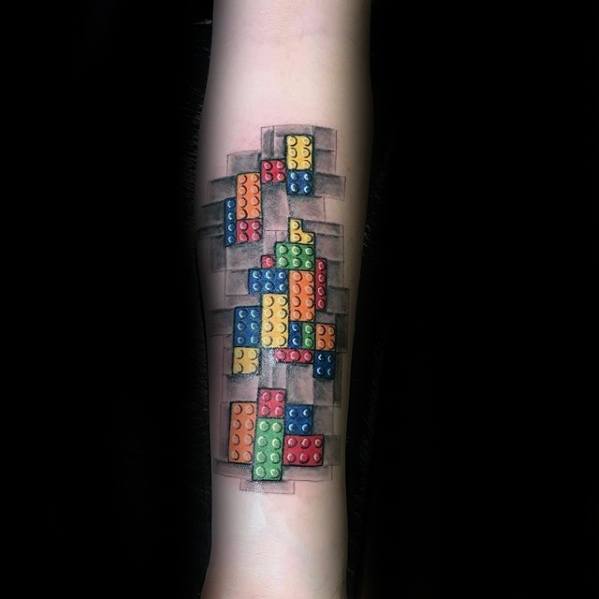Forearm tattoo of colorful Lego bricks appearing to break through the skin, with a 3D effect and shadowed background for depth.