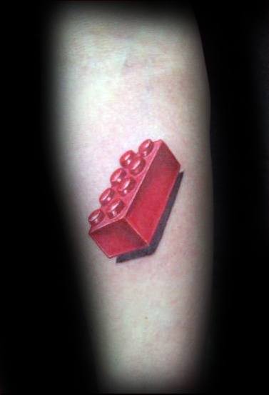 Realistic tattoo of a red Lego brick with bold shading and a dark shadow, creating a strong 3D effect on the forearm.