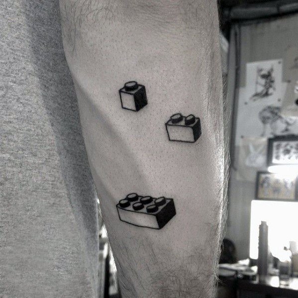 Minimal black and white tattoo of three Lego bricks in different sizes, arranged diagonally on the arm with simple shading.