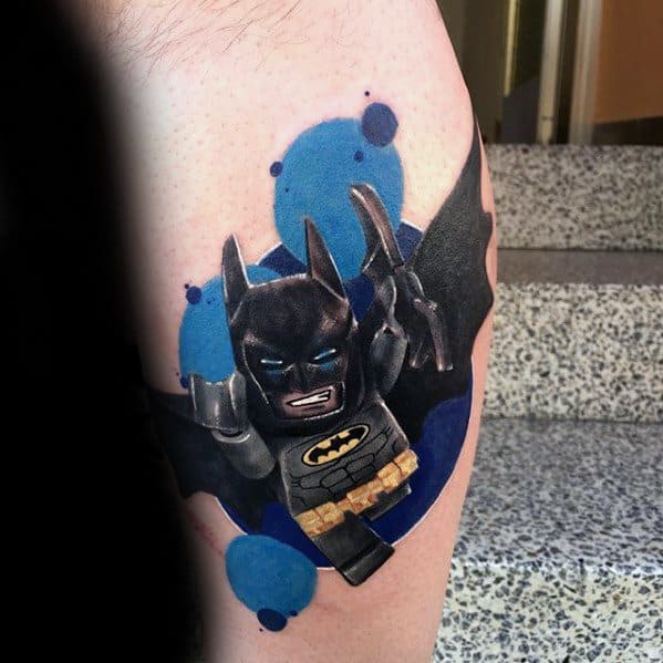 Dynamic tattoo of Lego Batman in mid-action with batarangs, set against a bold blue splash background for a comic-style effect.