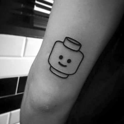 Simple black line tattoo of a classic Lego minifigure head with a smiling face, placed near the elbow for a playful minimalist look.