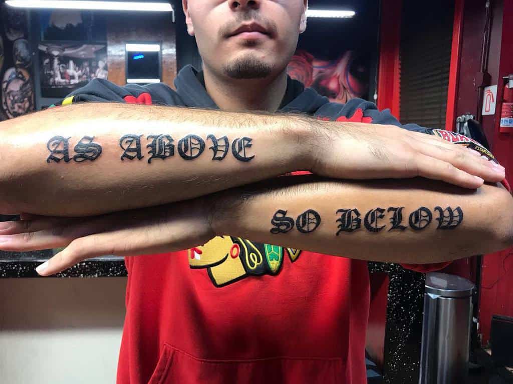 Forearm lettering tattoo showcasing 'As Above' and 'So Below' in bold gothic script, symbolizing universal balance and harmony