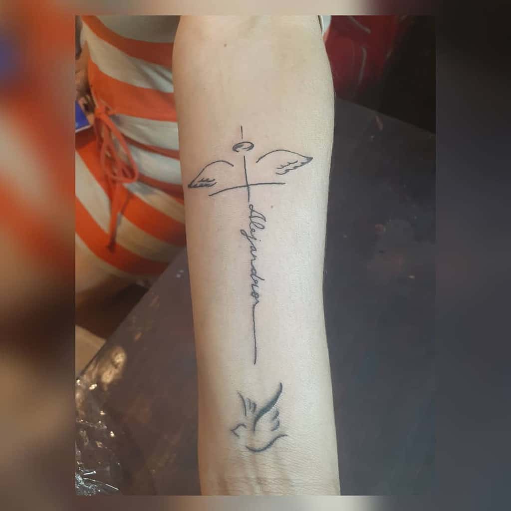 A forearm tattoo featuring a cross with wings, the name "Alejandro," and a small dove nestled below
