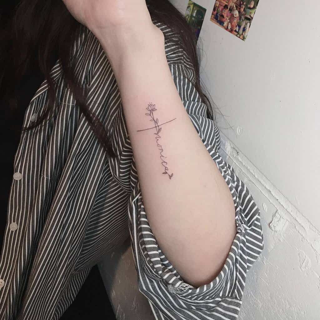 Forearm with a delicate tattoo of "monica" in cursive, a small heart intertwined with a tiny cross