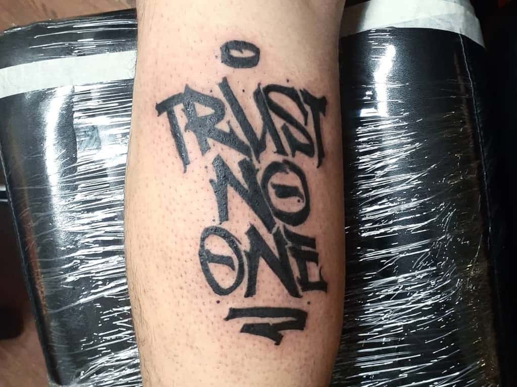 Black graffiti-style tattoo on an arm reading "TRUST NO ONE" in bold letters