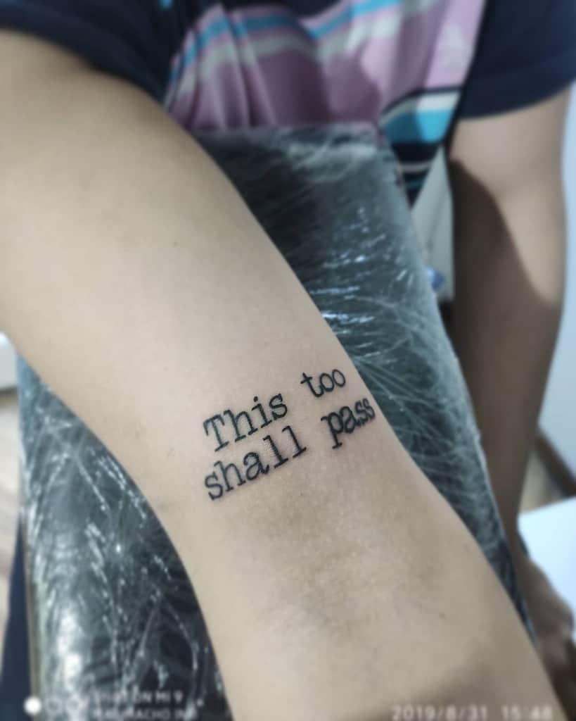 This image features a tattoo of the phrase "This too shall pass" on someone's forearm, presented in a typewriter-style font.