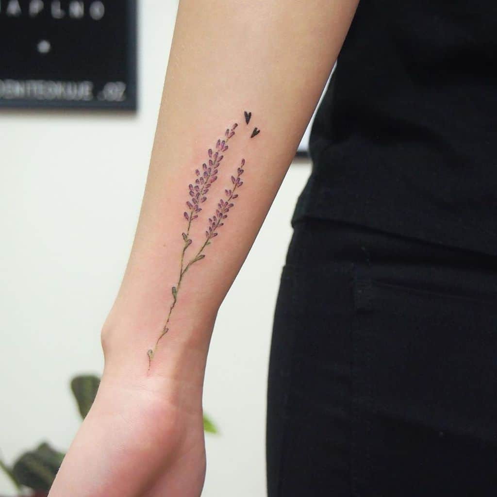 A minimalist lavender tattoo with delicate purple stems and small flying hearts near the top, placed on the inner arm.