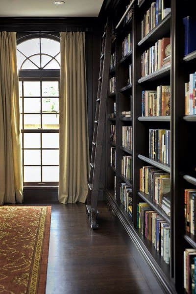 Library with tall bookshelves, a wooden ladder, and large window with curtains A patterned rug covers the dark floor