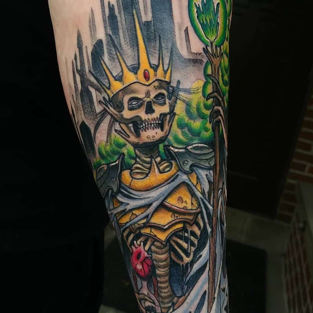 A vibrant tattoo of a skeletal lich wearing golden armor and a jeweled crown, holding a glowing green staff with a mystical background of a dark castle and green mist