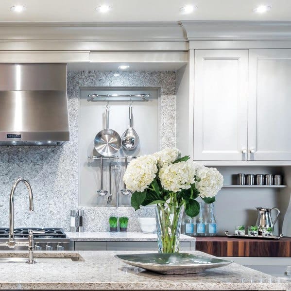 A modern kitchen featuring light grey cabinetry, crown molding, marble countertops, and stylish lighting with fresh flowers as a centerpiece