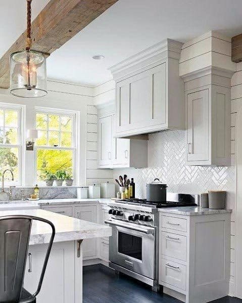 Light grey kitchen with white herringbone backsplash, marble countertops, and exposed wooden beam.