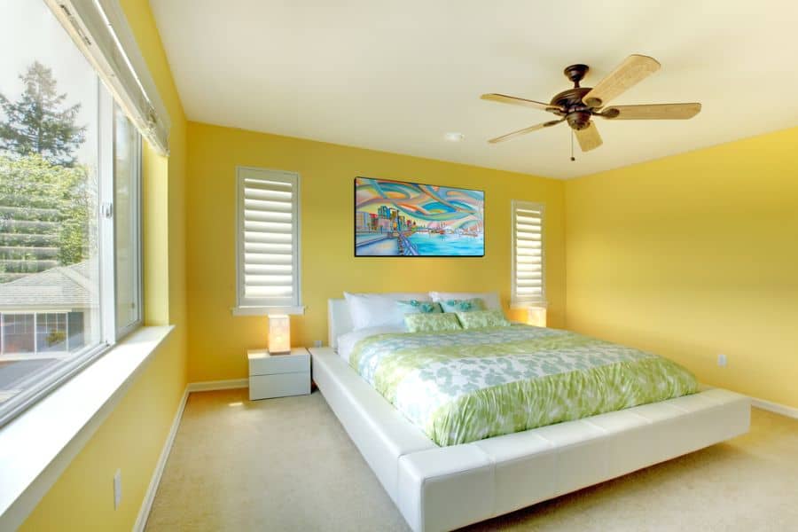 A vibrant bedroom with yellow walls, a ceiling fan, a large bed, and a colorful painting above it
