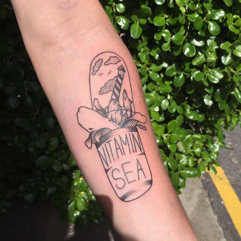 Creative tattoo of a pill capsule labeled 'Vitamin Sea,' bursting open to reveal ocean waves, shark, a lighthouse, and clouds