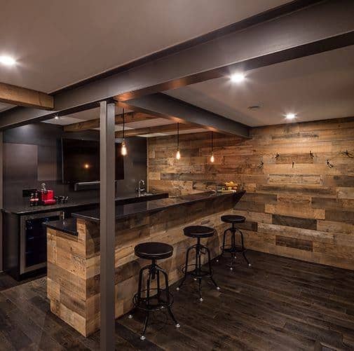 Rustic basement bar with wooden walls, industrial lighting, and dark wood flooring.