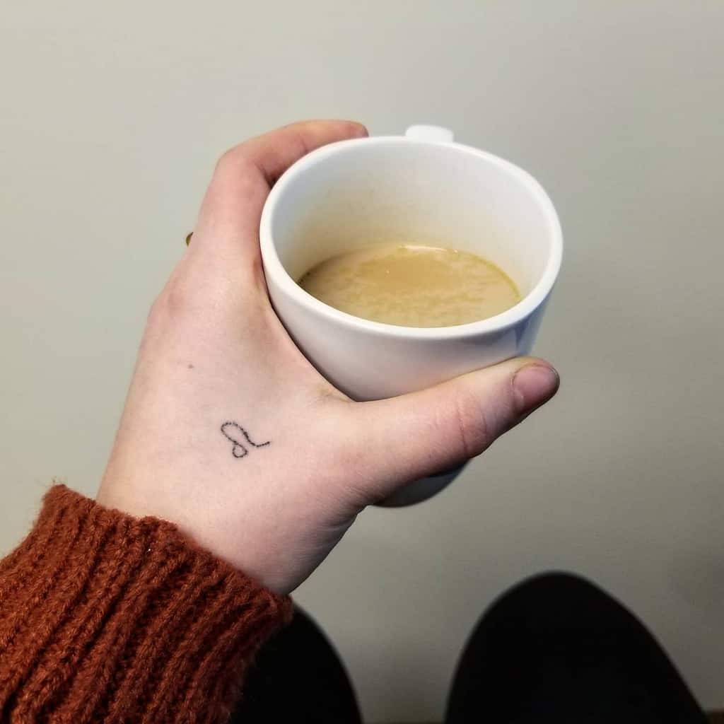 Minimalist black ink Leo tattoo on the hand, captured in a cozy moment with a warm drink.