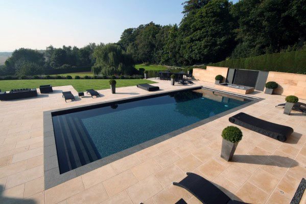 A sleek backyard pool featuring dark mosaic tiles, limestone decking, and lush green surroundings.